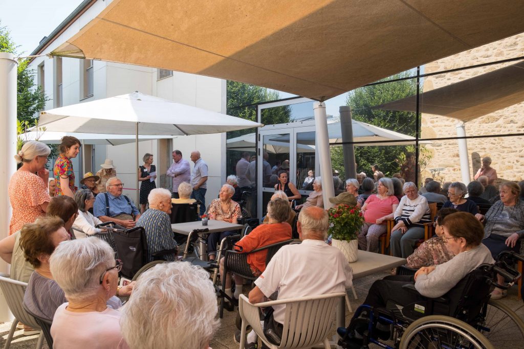 Reportage évènementiel en EHPAD. Résidents, professionnels et invités dans le patio de l'EHPAD, lors du vernissage d'une exposition photo.