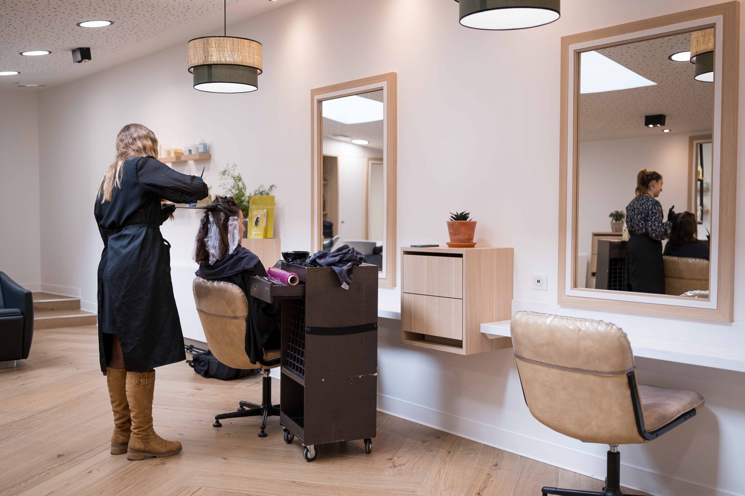 Vue du salon de coiffure, avec une coiffeuse coloriste réalisant une couleur à une cliente, avec le reflet de sa collègue dans le miroir